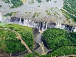 Szeroka panorama Wodospadów Wiktorii widziana z góry, z widocznym głębokim wąwozem, mgłą wodną i mostem łączącym Zambię z Zimbabwe pośród bujnej zieleni.