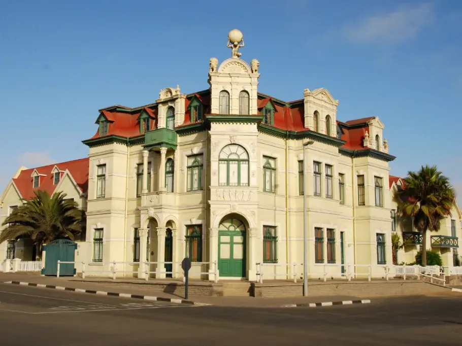 Zabytkowy budynek Hohenzollernhaus w mieście Swakopmund w Namibii. Jest to imponująca, narożna budowla w stylu neobarokowym o jasnokremowej elewacji i czerwonym dachu, na szczycie której znajduje się rzeźba Atlasa. Budynek otaczają palmy, charakterystyczne dla tego nadmorskiego kurortu.