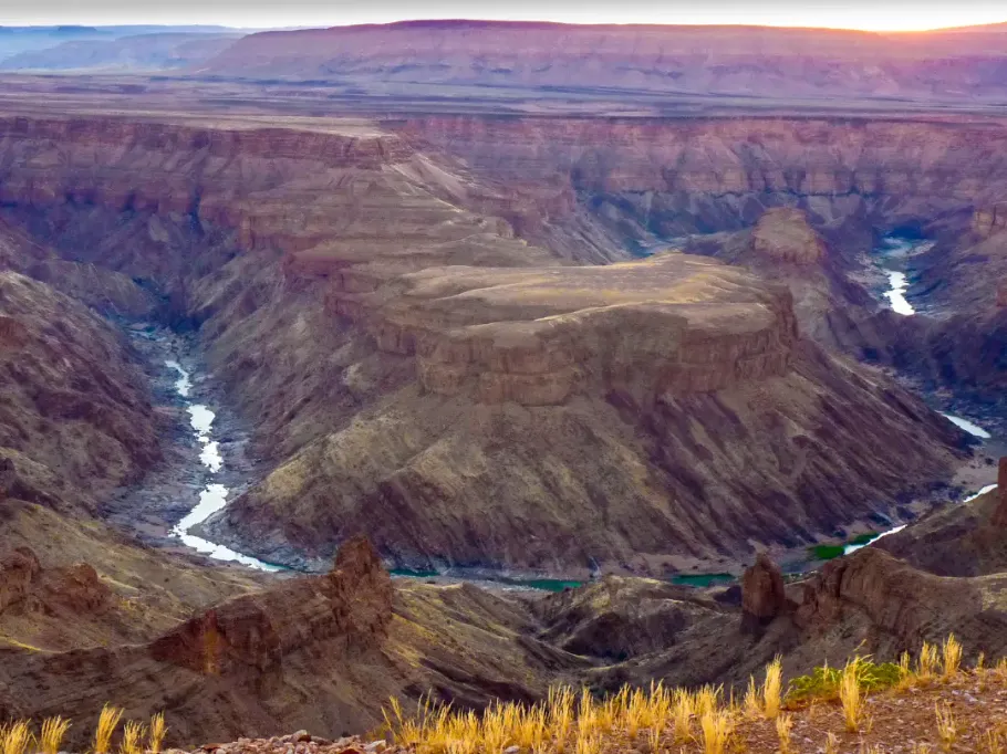 Szeroki, panoramiczny widok z góry na Kanion Fish River w Namibii. Rzeka wije się głęboko na dnie skalistego wąwozu o stromych, warstwowych zboczach w odcieniach brązu i czerwieni. Na pierwszym planie widać kępy suchej, żółtej trawy, a w tle rozciąga się zamglony płaskowyż.