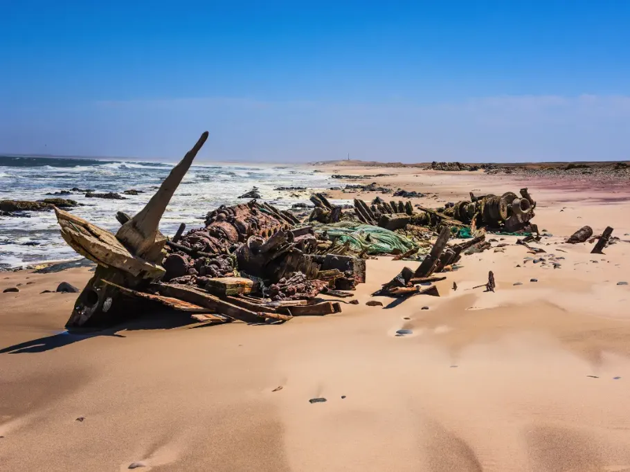 Widok z lotu ptaka na spektakularne wybrzeże, gdzie ogromne, złociste wydmy pustyni bezpośrednio spotykają się z ciemnoniebieskim oceanem. Po lewej stronie widać wzburzone wody z białymi grzywami fal uderzającymi o brzeg. Po prawej rozciąga się pofalowane morze piasku. Wzdłuż wąskiego pasa plaży widoczne są drobne ślady kół samochodów terenowych, co podkreśla ogromną skalę krajobrazu. Sandwich Harbour, Namibia