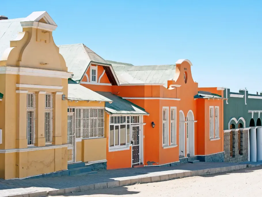Rząd kolorowych, zabytkowych budynków w stylu niemieckiego kolonializmu w Lüderitz w Namibii. W pełnym słońcu widać przylegające do siebie domy o jaskrawych elewacjach: żółtej, intensywnie pomarańczowej i zielonej. Budynki mają charakterystyczne, ozdobne szczyty dachowe, białe obramowania okien i spadziste dachy, a nad nimi rozciąga się bezchmurne, błękitne niebo.