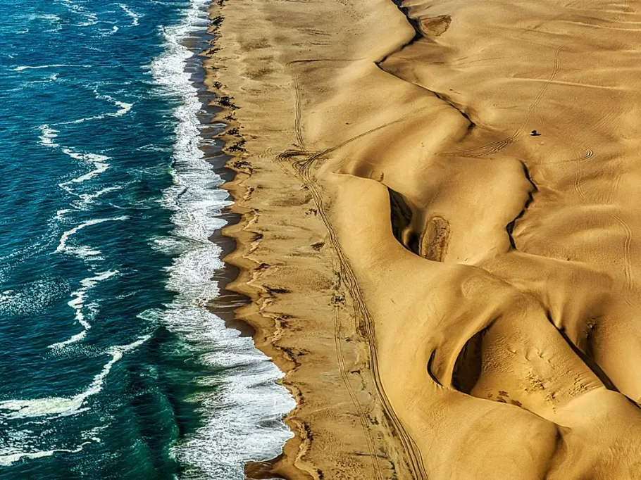 Widok z lotu ptaka na spektakularne wybrzeże, gdzie ogromne, złociste wydmy pustyni bezpośrednio spotykają się z ciemnoniebieskim oceanem. Po lewej stronie widać wzburzone wody z białymi grzywami fal uderzającymi o brzeg. Po prawej rozciąga się pofalowane morze piasku. Wzdłuż wąskiego pasa plaży widoczne są drobne ślady kół samochodów terenowych, co podkreśla ogromną skalę krajobrazu. Sandwich Harbour, Namibia
