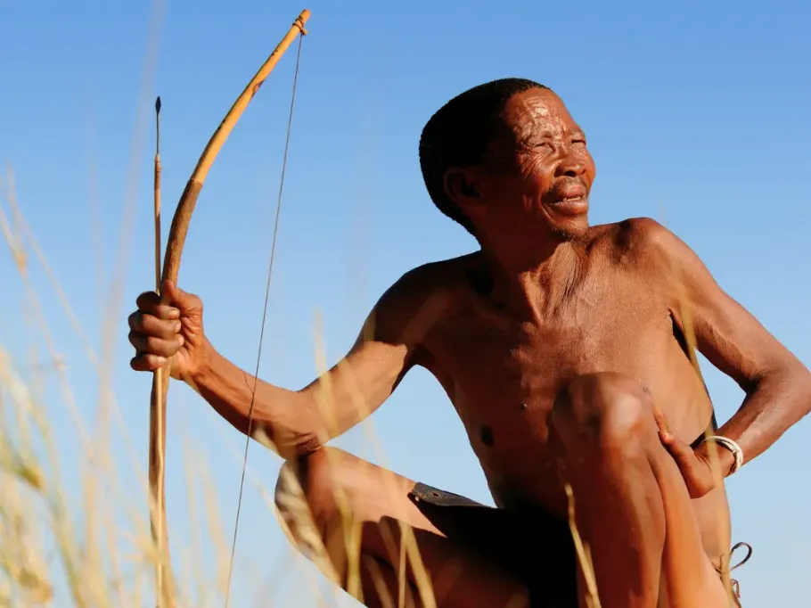 Starszy myśliwy z ludu San w Namibii kuca pośród wysokich, suchych traw pod błękitnym niebem, trzymając w ręku tradycyjny drewniany łuk i strzałę i spoglądając w dal.