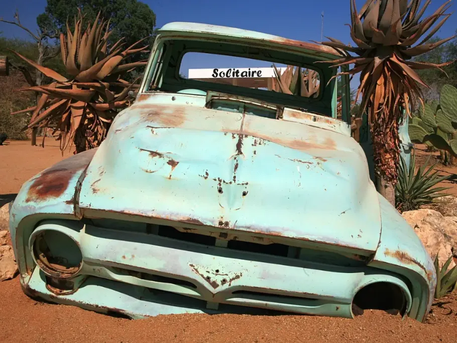 Malownicze cmentarzysko starych, zardzewiałych samochodów z połowy XX wieku na tle pustynnego krajobrazu Solitaire w Namibii. Wraki o rudej, spalonej słońcem karoserii są częściowo wkopane w piasek i otoczone kępami suchych traw oraz kaktusami. W tle widać budynek stacji benzynowej i sklepu utrzymany w stylu Dzikiego Zachodu.