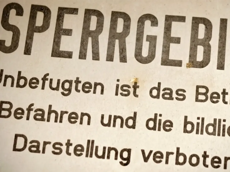 Zbliżenie na stary, zniszczony znak z dużym, czarnym napisem 'SPERRGEBIET' u góry. Poniżej widoczne są fragmenty niemieckiego tekstu: 'Unbefugten ist das...', 'Befahren und die bildli...' oraz 'Darstellung verboten', ostrzegające przed wstępem i zakazem fotografowania na beżowym, wyblakłym tle.
