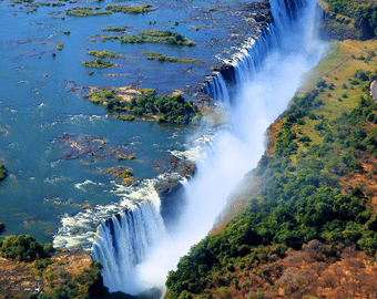 Szeroka panorama Wodospadów Wiktorii widziana z góry, z widocznym głębokim wąwozem, mgłą wodną i mostem łączącym Zambię z Zimbabwe pośród bujnej zieleni.