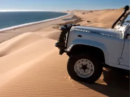 Widok z boku na przód białego samochodu terenowego 4x4 (typu Land Rover Defender) z podwyższonym wlotem powietrza, stojącego na krawędzi wysokiej piaszczystej wydmy. W dole rozciąga się wąski pas plaży, gdzie pustynia Namib styka się z ciemnoniebieskimi wodami Oceanu Atlantyckiego pod bezchmurnym niebem.